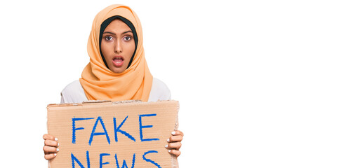 Young brunette arab woman holding fake news banner scared and amazed with open mouth for surprise,...