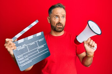 Handsome middle age man holding video film clapboard and megaphone in shock face, looking skeptical...