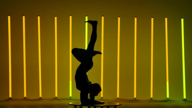The athlete performs gymnastics against the backdrop of neon lights. The girl performs a handstand, on the chest, demonstrates the movements of her legs in the air. Dark silhouette. Slow motion.