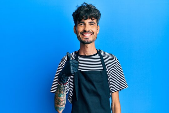 Young hispanic man wearing barber apron smiling happy and positive, thumb up doing excellent and approval sign