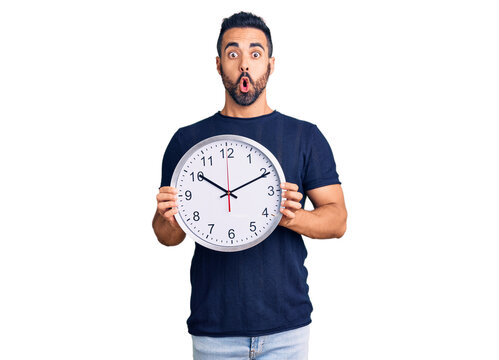 Young hispanic man holding big clock scared and amazed with open mouth for surprise, disbelief face