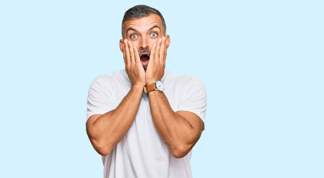 Middle age handsome man wearing casual white tshirt afraid and shocked, surprise and amazed expression with hands on face