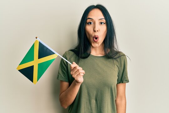 Young hispanic girl holding jamaica flag scared and amazed with open mouth for surprise, disbelief face