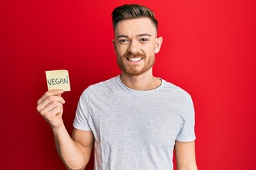 Young redhead man holding sticker with vegan word looking positive and happy standing and smiling with a confident smile showing teeth