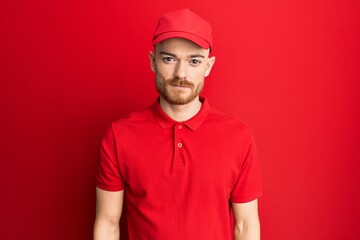 Young redhead man wearing delivery uniform and cap with serious expression on face. simple and natural looking at the camera.