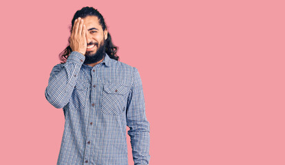 Young arab man wearing casual clothes covering one eye with hand, confident smile on face and surprise emotion.