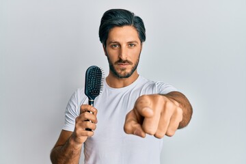 Young hispanic man styling hair using comb pointing with finger to the camera and to you, confident gesture looking serious