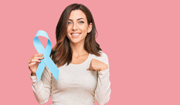 Young brunette woman holding blue ribbon pointing finger to one self smiling happy and proud