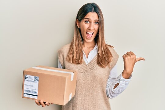 Brunette young woman holding delivery package pointing thumb up to the side smiling happy with open mouth