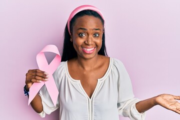 Young african woman holding pink cancer ribbon celebrating achievement with happy smile and winner...