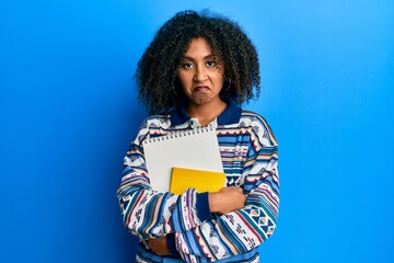 Beautiful african american woman with afro hair holding books skeptic and nervous, frowning upset...