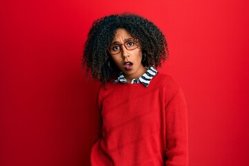 Beautiful african american woman with afro hair wearing sweater and glasses in shock face, looking skeptical and sarcastic, surprised with open mouth