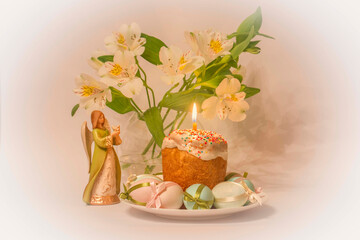 Easter. Pastry decorated with colored beads and a burning candle. Traditional colored eggs decorated with ribbons. There is an angel figurine on the table. In the background, a vase with flowers.