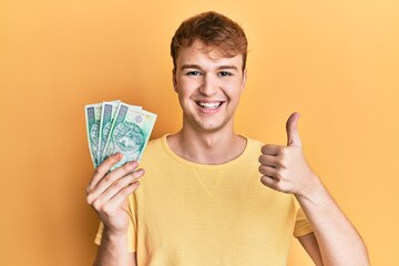Young caucasian man holding 50 polish zloty banknotes smiling happy and positive, thumb up doing...