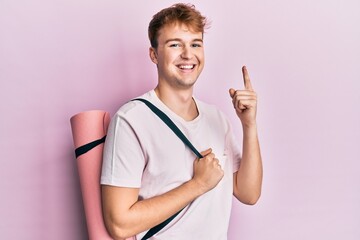 Young caucasian man holding yoga mat smiling with an idea or question pointing finger with happy face, number one