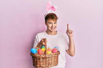 Young caucasian man wearing cute easter bunny ears holding wicker basket with colored eggs surprised with an idea or question pointing finger with happy face, number one
