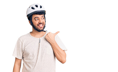 Young hispanic man wearing bike helmet smiling with happy face looking and pointing to the side with thumb up.