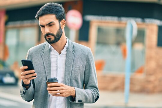 Young hispanic businessman with serious expression using smartphone and drinking coffee at the city.