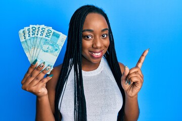 Young african american woman holding 100 brazilian real banknotes smiling happy pointing with hand and finger to the side