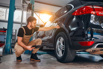 Handsome mechanic in uniform is making notes examining car in auto service. - obrazy, fototapety, plakaty