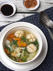 Vegetable clear soup with chicken meat balls served in white bowl on  crochet place mat along with soy sauce and fried shallot on white rustic wooden table. Healthy food. Selective focus. 