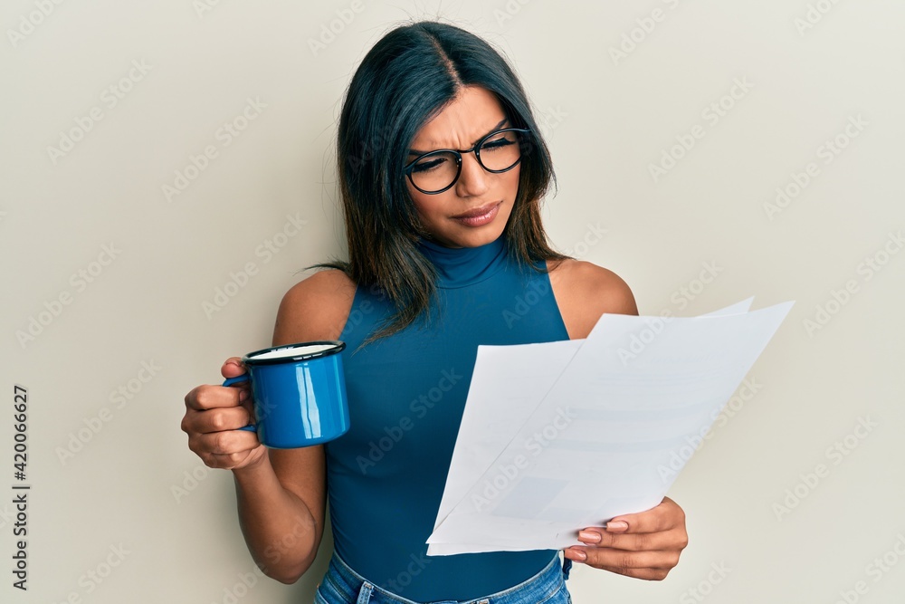 Wall mural Young latin transsexual transgender woman drinking cup of coffee and reading paper clueless and confused expression. doubt concept. - Wall murals