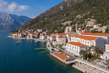 Fototapeta na wymiar Aerial shot of the old coastal town of Perast at the foot of the mountain. Seaside promenade, residential buildings with traditional balkan red roofs, ancient Cathedral and coastline