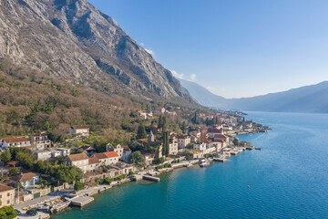 Naklejka na ściany i meble Aerial shot of the old coastal town of Perast at the foot of the mountain. Seaside promenade, residential buildings with traditional balkan red roofs, ancient Cathedral and coastline