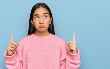 Young asian woman wearing casual winter sweater pointing up looking sad and upset, indicating...