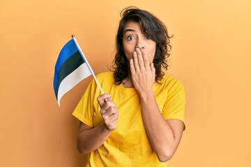 Young hispanic woman holding estonia flag covering mouth with hand, shocked and afraid for mistake. surprised expression
