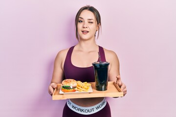 Young hispanic girl wearing sportswear eating a tasty classic burger with fries and soda winking looking at the camera with sexy expression, cheerful and happy face.