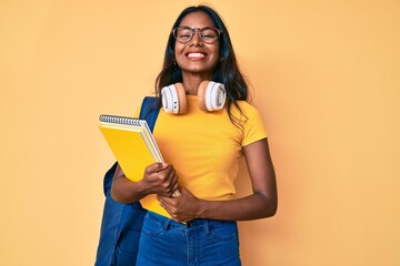 Young indian girl holding student backpack and books looking positive and happy standing and smiling with a confident smile showing teeth