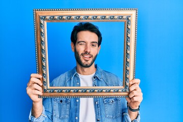 Young hispanic man holding empty frame winking looking at the camera with sexy expression, cheerful...