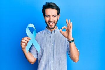 Fototapeta premium Young hispanic man holding blue ribbon doing ok sign with fingers, smiling friendly gesturing excellent symbol