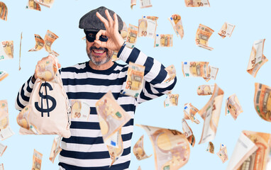 Middle age handsome man wearing burglar mask holding money bag smiling happy doing ok sign with hand on eye looking through fingers