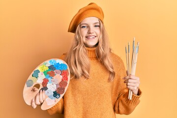 Beautiful young caucasian girl holding paintbrush and painter palette smiling with a happy and cool smile on face. showing teeth.