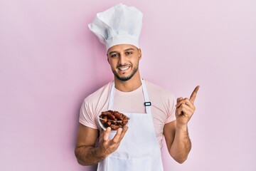 Young arab man wearing professional cook uniform holding bowl with dates smiling happy pointing with hand and finger to the side