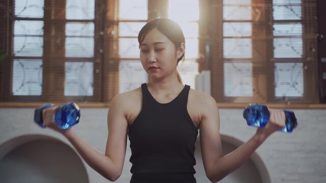 Young Asian female using laptop watching tutorial online at home for exercise and workout with dumbbell in living room during lockdown. New normal healthy woman fitness at apartment during gym close