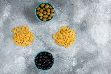 Uncooked farfalle and bowls of olives on marble background