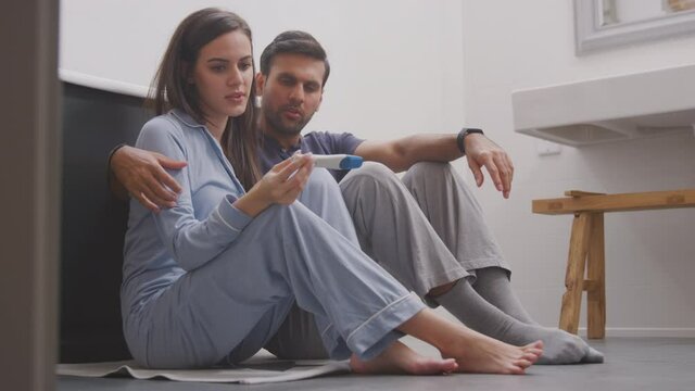 Disappointed couple wearing pyjamas sitting on floor of bathroom at home looking sad after negative result from home pregnancy test - shot in slow motion