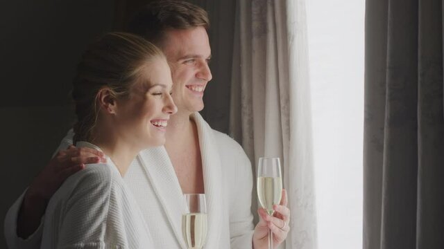 Relaxed couple wearing bathrobes on hotel or spa break standing by open curtains making a toast with champagne- shot in slow motion