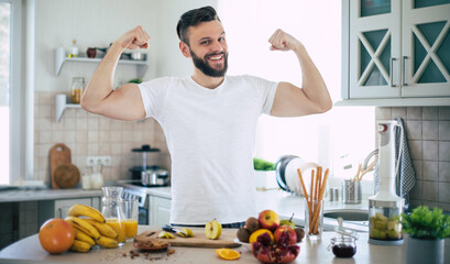 Handsome young sporty smiling man in the kitchen is preparing vegan healthy fruits salad and smoothie in a good mood