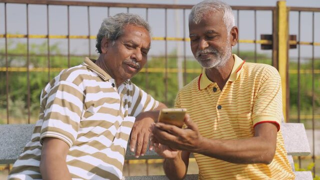 senior man showing mobile phone to his friend while sitting at park - Elderly people watching online news or entertainment on smartphone - concept of old men using social media,internet and technology