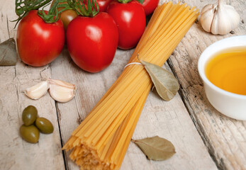 Raw spaghetti pasta next to fresh red tomatoes on a branch, garlic, olive oil and bay leaves on a wooden surface.