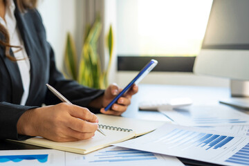 Young businesswoman sitting at the table and taking notes in a notebook. On the table is a laptop, smartphone. On computer screen graphics and charts. Student learning online. Blogger.