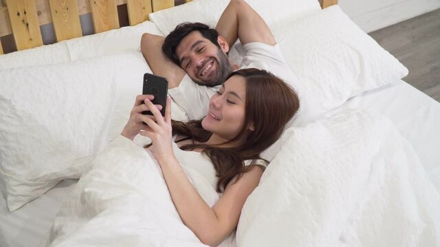 Footage b roll of Couple using smart phone and happy on bed. Beautiful couple sitting on the bed and looking photo from the trip on smartphone, touching screen, Watching movie or chatting app.