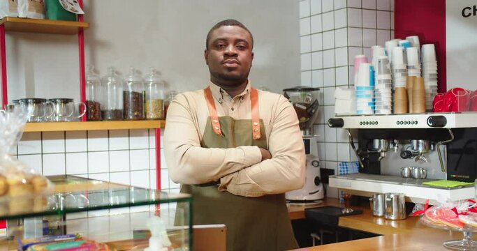 Portrait of professional barista in modern coffee shop with minimalist interior. Young Afro American guy standing beding bar, looking at camera and crossing arms. Cafe, business concept.