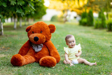 Cute little girl sitting on green grass with big Teddy bear in yellow summer dress in summer, a place for text
