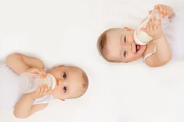 Fototapeten twin babies boy and girl with a bottle of milk on a white bed at home, baby food concept, place for text © Any Grant
