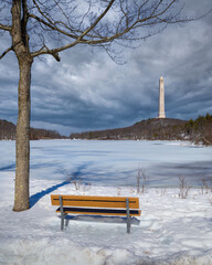 Empty bench looking across Lake Marcia towards the New Jersey High Point Monument on a snow-covered winter day